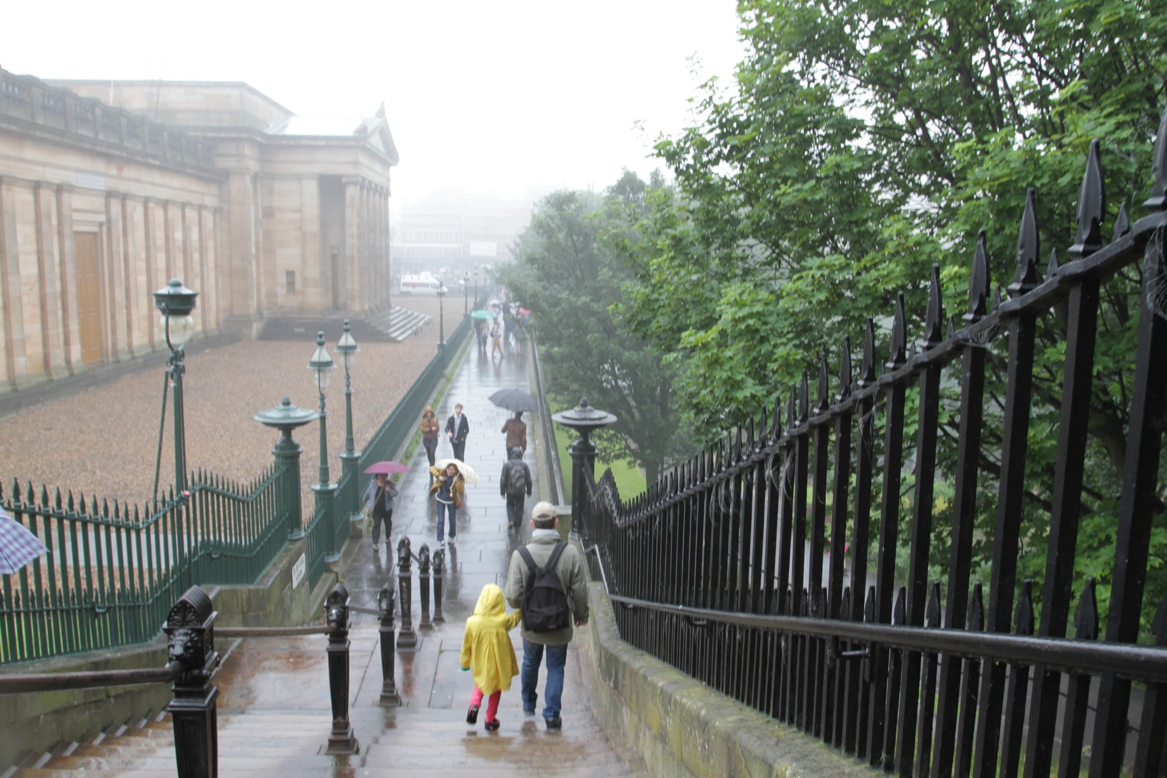 A rainy street scene with a Neoclassical building and pedestrians under umbrellas, straight out of camera.
